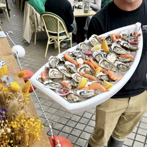 Écailler de Dimanch'Huîtres portant un grand présentoir en forme de bateau blanc garni d'huîtres ouvertes, de crevettes roses fraîches, de citrons et de sauces. Prestation traiteur en terrasse extérieure en Gironde.