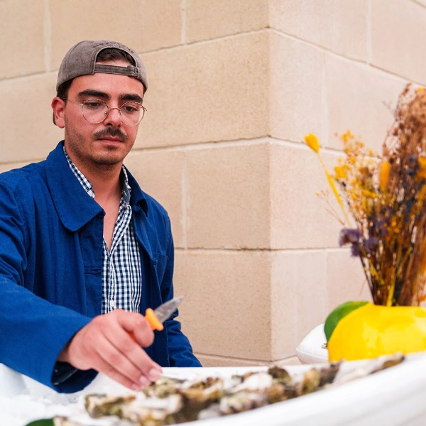 Écailler de Dimanch'Huîtres portant un grand présentoir en forme de bateau blanc garni d'huîtres ouvertes, de crevettes roses fraîches, de citrons et de sauces. Prestation traiteur en terrasse extérieure en Gironde.
