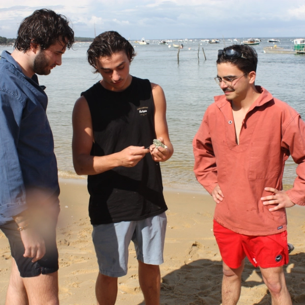 Trois membres de l'équipe Dimanch'Huîtres debout sur une plage de sable fin en Gironde, observant attentivement une huître fraîche tenue dans la main au bord du Bassin d'Arcachon avec des bateaux en arrière-plan.