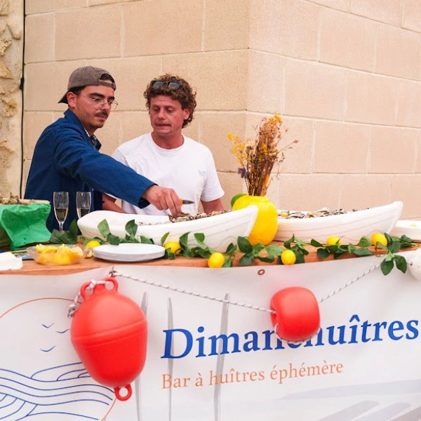 Deux membres de l'équipe de Dimanch'Huîtres en train d'ouvrir des huîtres fraîches derrière un buffet décoré de citrons, de bouées rouges et de barques blanches remplies de glace. Animation écailler professionnelle lors d'un événement corporate à Bordeaux.