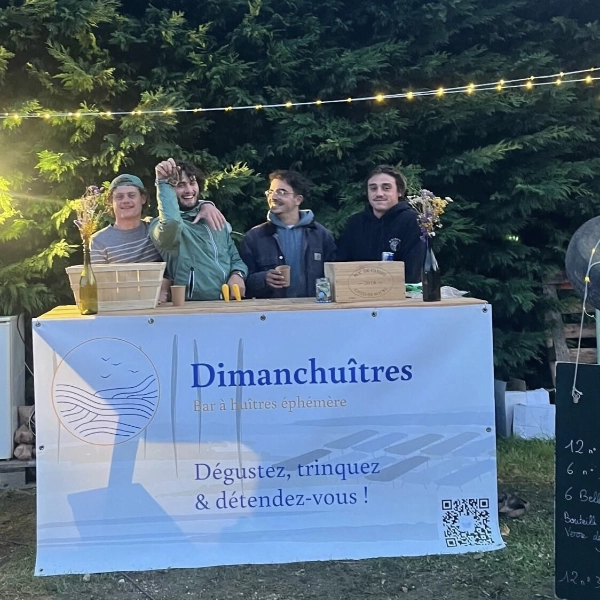 Les quatre membres souriants de l'équipe Dimanch'Huîtres derrière leur comptoir en bois officiel orné d'une bâche publicitaire avec le slogan Dégustez, trinquez et détendez-vous, lors du festival L'Ode Mère en Gironde.