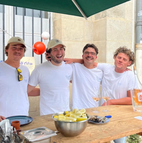 L'équipe d'écaillers professionnels de Dimanch'Huîtres souriant derrière leur bar en bois lors d'un événement haut de gamme en Gironde. Buffet traiteur avec huîtres fraîches, citrons et dégustation de vin devant une bâtisse en pierre typique de la région de Bordeaux.